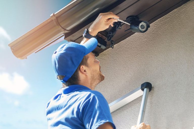 A man wearing a blue shirt is repairing a camera mounted on the side of a house.